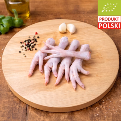 Łapki z kurczaka ekologicznego — cztery surowe łapki drobiowe ułożone na drewnianej desce z ząbkami czosnku, kolorowym pieprzem i oliwą w tle
