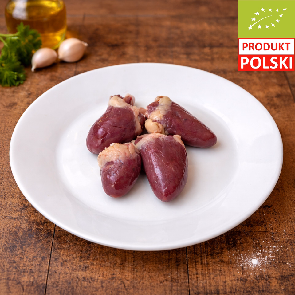 Serca z kurczaka pastwiskowego — cztery surowe serca drobiowe na białym talerzu z ząbkami czosnku, świeżą natką pietruszki i oliwą w tle