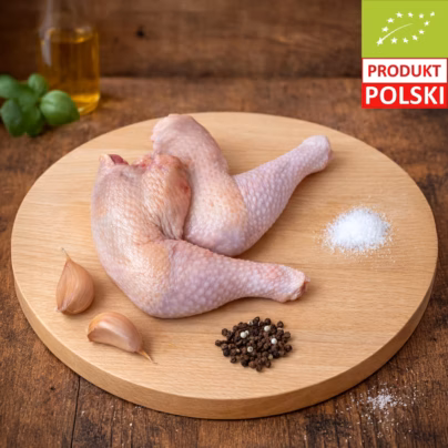 Udka z kurczaka ekologicznego — dwie surowe nogi z kurczaka pastwiskowego ze skórą ułożone na drewnianej desce z czosnkiem, kolorowym pieprzem, solą gruboziarnistą i bazylią w tle