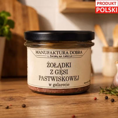 Żołądki z gęsi pastwiskowej w galarecie w szklanym słoiku z etykietą Manufaktura Dobra, na drewnianym blacie z czosnkiem i ziarnami pieprzu w tle